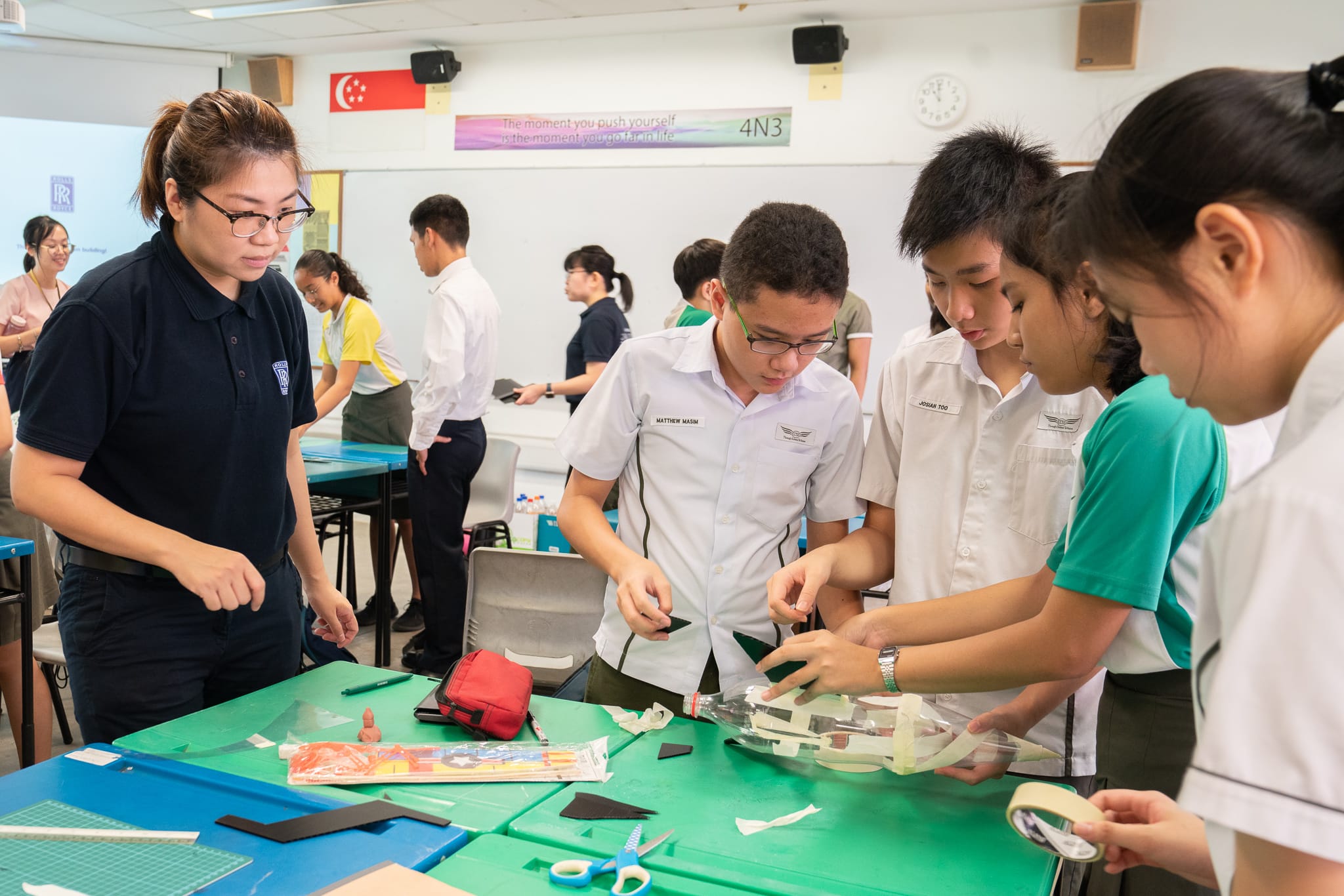 Students in school uniforms work on a hands-on craft project at a green table in a Singapore classroom.